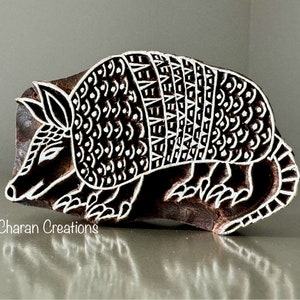 May include: A wooden block carving of an armadillo with intricate details. The armadillo is carved in a detailed, stylized design with a patterned shell. The carving is a dark brown color and has a white outline.