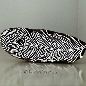 Puede incluir: Un sello de impresión de madera en blanco y negro con un diseño detallado de una pluma de pavo real. El sello está tallado con líneas intrincadas y un patrón de ojo circular.