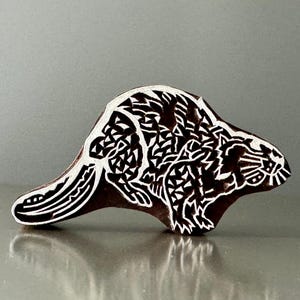 May include: A dark brown wooden printing block carved into the shape of a beaver. The surface features intricate white geometric patterns and lines that define the animal's body, tail, and facial features, creating a high-contrast relief design.