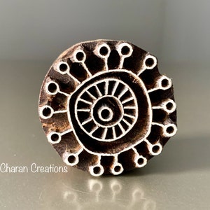 May include: A wooden block printing stamp with a circular design. The design features a central circle with radiating lines and a smaller circle inside. The outer edge of the stamp is decorated with a series of small circles.