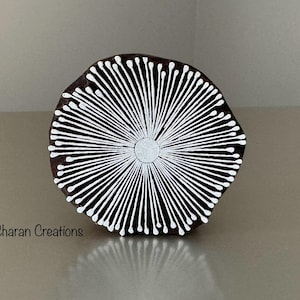 May include: A round, wooden printing block with a radial design of white lines emanating from a central circle. The block is dark brown with a slightly irregular edge. The text "© Charan Creations" is visible.