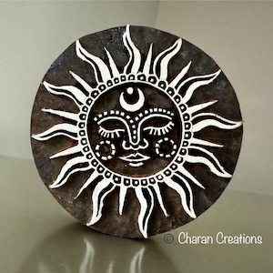 May include: A wooden block printing stamp with a black and white design of a sun with a face and a crescent moon. The stamp is round and has a brown wooden surface.