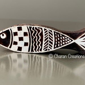 Könnte beinhalten: Ein holzgeschnitzter Fischblock mit weißen geometrischen Mustern. Der dunkelbraune Block hat ein detailliertes Design aus Quadraten, Wellen und Dreiecken. Der Text "© Charan Creations" ist auf der Seite sichtbar.