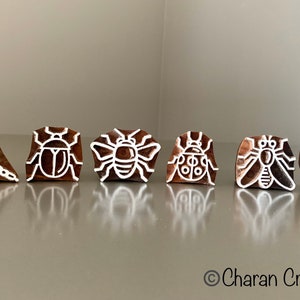 Könnte beinhalten: Satz mit sechs Holzstempeln mit verschiedenen Insektenmotiven, darunter eine Libelle, ein Käfer, eine Biene, ein Marienkäfer, eine Fliege und eine Ameise. Die Stempel sind aus Holz geschnitzt und haben weiße Umrisse.