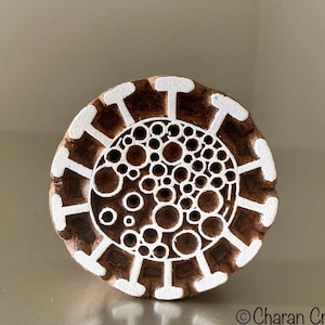 Puede incluir: Un bloque de madera redondo con base marrón y diseño pintado en blanco. El diseño presenta un círculo central lleno de círculos más pequeños, rodeado de formas blancas. Probablemente utilizado para imprimir o estampar.