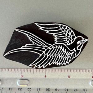 May include: A wooden block printing stamp with a white design of a stylized bird with wings spread. The bird is facing right and has a long tail.