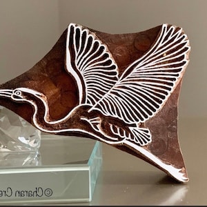 May include: A wooden block featuring a carved design of a bird in flight. The bird is white against a dark brown background. The block is rectangular with a curved edge.