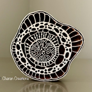 Puede incluir: Un sello de impresión de madera en blanco y negro con un diseño circular que presenta intrincados patrones y remolinos. El sello está tallado con un diseño tipo mandala.