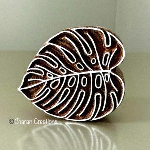 May include: A brown wooden stamp in the shape of a monstera leaf, featuring white detailed veins and cutouts. The stamp is designed for block printing and has the text "© Charan Creations" on the bottom left.