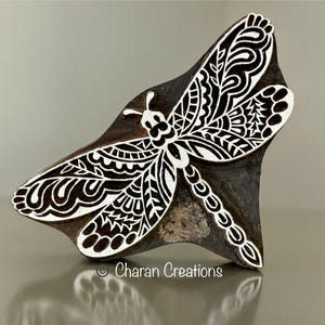 Könnte beinhalten: Eine Holzschnitzerei einer Libelle mit filigranen Details. Die Libelle ist in einem schwarz-weißen Muster geschnitzt und auf einem braunen Holzgrund platziert.