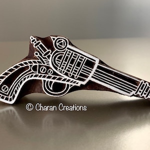May include: A white wooden block carved with a detailed design of a pistol. The block is brown and has a white outline of the pistol. The text "Charan Creations" is visible at the bottom of the image.