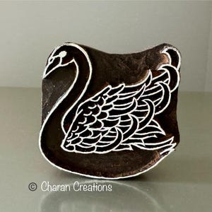 May include: A dark brown wooden block featuring a white swan design. The swan has a curved neck and detailed wings. The block is likely used for printing or stamping. The design is a simple, elegant representation of a swan.