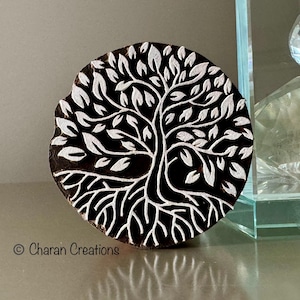 May include: Round, wooden block with a tree of life design. The tree is depicted with white leaves and branches against a dark brown background. The block has a slightly irregular edge. The text "© Charan Creations" is at the bottom.