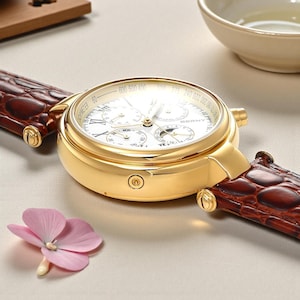 May include: A gold-toned wristwatch with a white face and brown leather band. The watch face features multiple sub-dials and gold hands. The band has a crocodile texture. A pink flower is in the foreground.