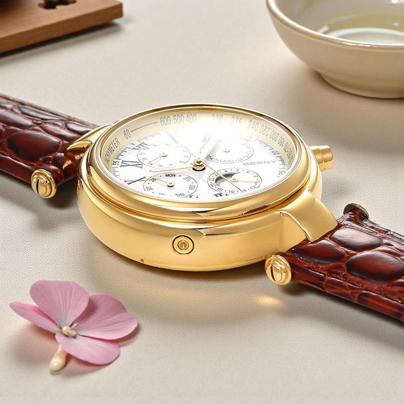 May include: A gold-toned wristwatch with a white face and brown leather band. The watch face features multiple sub-dials and gold hands. The band has a crocodile texture. A pink flower is in the foreground.