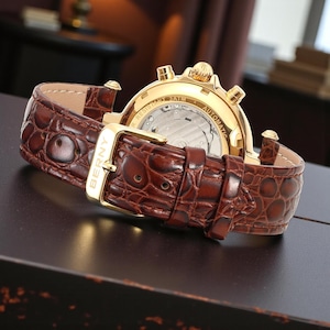 May include: A gold-toned wristwatch with a brown leather band. The watch features a textured band with a gold buckle. The watch face is visible, displaying the inner workings. The watch is labeled "BERNY" on the buckle.