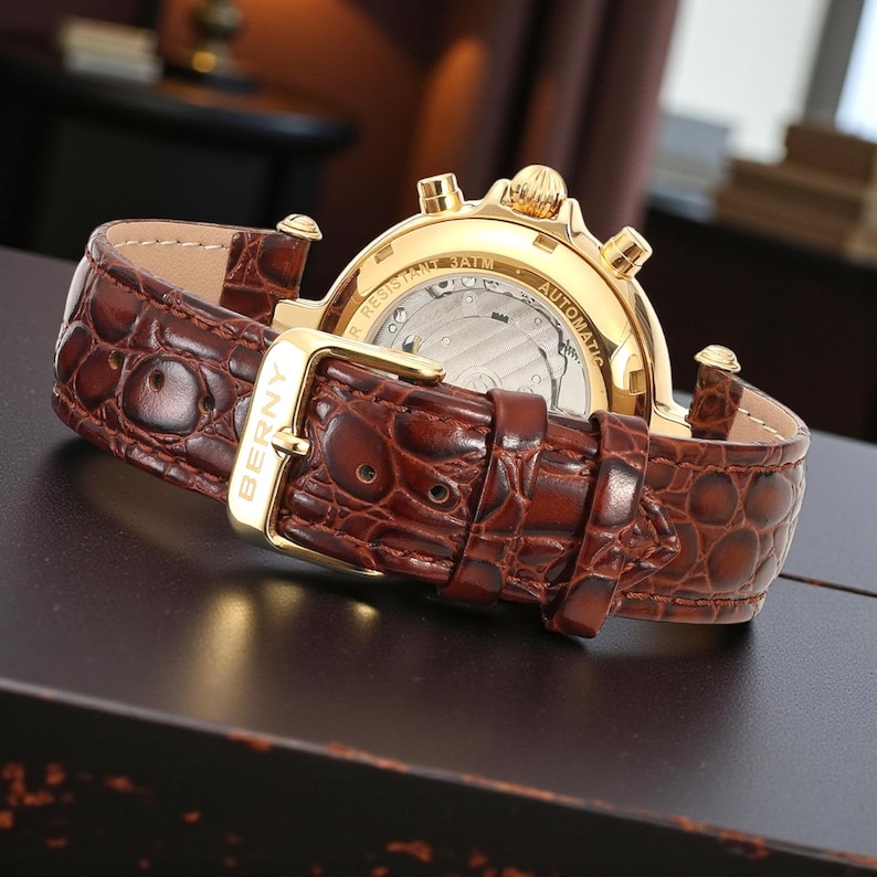 May include: A gold-toned wristwatch with a brown leather band. The watch features a textured band with a gold buckle. The watch face is visible, displaying the inner workings. The watch is labeled "BERNY" on the buckle.