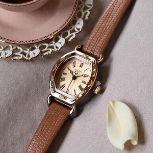 Puede incluir: Un reloj de pulsera dorado con una correa de cuero marrón. La esfera del reloj es dorada con números romanos negros y la marca "JULIUS". La correa tiene un patrón texturizado similar a la piel de reptil.