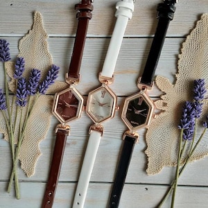 Puede incluir: Tres relojes de mujer con esferas hexagonales y correas finas de cuero en marrón, blanco y negro. Las esferas tienen un bisel color oro rosa y un diseño minimalista. Los relojes se exhiben sobre una superficie de madera clara.