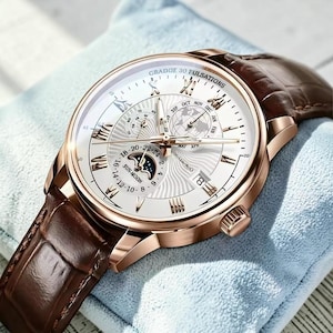May include: A wristwatch with a rose gold-tone case, a white dial featuring Roman numerals, and a brown leather strap. The dial displays a world map, a sun-moon phase indicator, a date window, and the text GRADDE 30 PULSATIONS.
