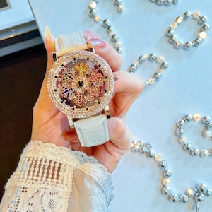 Könnte beinhalten: Eine goldfarbene Armbanduhr mit weißem Lederarmband. Das Zifferblatt der Uhr ist mit funkelnden Kristallen und einem farbenfrohen, verzierten Design besetzt. Die Uhr wird vor einem hellblauen Hintergrund gehalten.