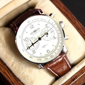 May include: A Zeppelin chronograph wristwatch with a white dial, black numerals, and three sub-dials. The watch features a tachymeter scale, silver-tone hands, and a brown leather strap with a crocodile-embossed texture, presented in a wooden box.