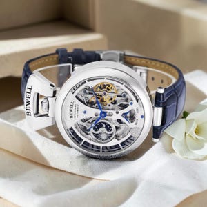 Puede incluir: Un reloj de pulsera automático plateado con una correa de cuero azul. La esfera del reloj está abierta, revelando el mecanismo interno. El reloj tiene la marca "BEWELL" en el cierre. El reloj está sobre una superficie blanca.