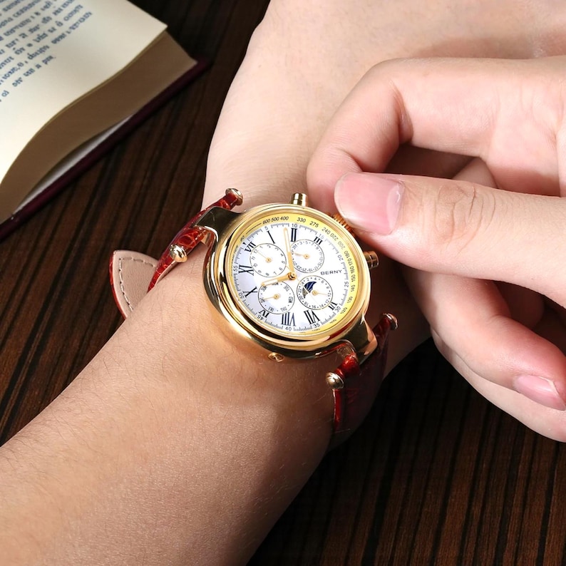 May include: A gold-toned wristwatch with a white face and multiple sub-dials. The watch has Roman numeral hour markers and a red leather band. The watch is approximately 1.5 inches in diameter.