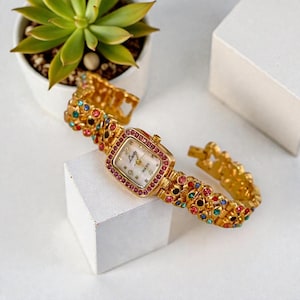 May include: Gold-toned quartz watch with a square face and a band adorned with colorful gemstones. The watch face is white with black numerals and the word "Lady" in script. The watch is displayed on a white surface.