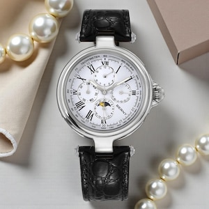 May include: A silver-toned wristwatch with a white face, featuring multiple subdials and Roman numeral hour markers. The watch has a black leather band and a tachymeter scale. The watch is surrounded by a pearl necklace and a cream-colored fabric.