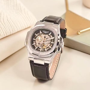 Könnte beinhalten: Eine silberfarbene Armbanduhr mit skelettiertem Zifferblatt, das die internen mechanischen Komponenten freilegt. Die Uhr verfügt über ein schwarzes Lederarmband mit strukturierter Oberfläche und den Markennamen LONGLUX in Weiß.
