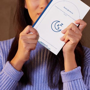 May include: A white Sleep Journal with blue text and a crescent moon and ZZZ graphic. The journal is held by a person wearing a light blue sweater. The journal's cover reads "Sleep Journal" and includes the text "A safe space to track your sleep, understand your patterns, and unlock..."