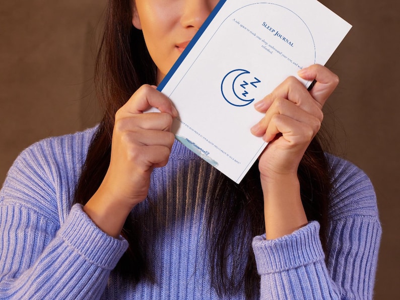 May include: A white Sleep Journal with blue text and a crescent moon and ZZZ graphic. The journal is held by a person wearing a light blue sweater. The journal's cover reads "Sleep Journal" and includes the text "A safe space to track your sleep, understand your patterns, and unlock..."