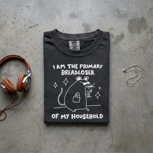 May include: Dark gray t-shirt with the text "I AM THE PRIMARY BREADLOSER OF MY HOUSEHOLD" and a cartoon rat wearing sunglasses and holding a cocktail. The shirt is folded on a gray surface, with headphones and a necklace nearby.