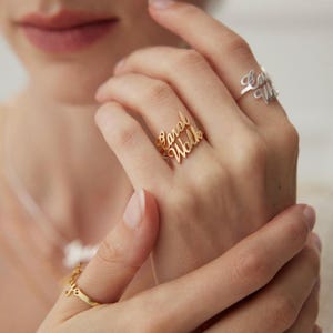 Può includere: Due anelli con scritte personalizzate in corsivo. Un anello è color oro e mostra il nome Carol Walker, mentre l'altro è color argento. Un terzo anello color oro con testo in corsivo è visibile sul dito inferiore.