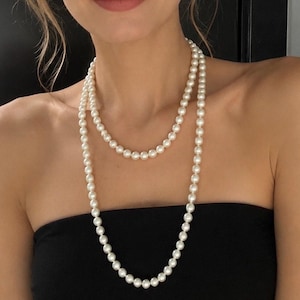 Puede incluir: Un collar largo de doble hebra con perlas redondas y uniformes de color blanco roto. El collar se lleva en dos vueltas alrededor del cuello, y las perlas presentan un acabado liso y brillante con un tamaño constante en toda la longitud del hilo.