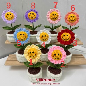 May include: A collection of handmade crochet flowers in various colors, each with a smiling face center. The flowers are potted in white containers and arranged on a wooden shelf. The flowers include yellow, pink, red, orange, blue, purple and white.