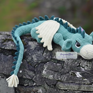 May include: A crocheted dragon toy in shades of teal and cream, resting on a stone surface. The dragon features detailed scales, wings, and a friendly expression. The tail ends in a tassel. The text "HungmmoStore" is visible.