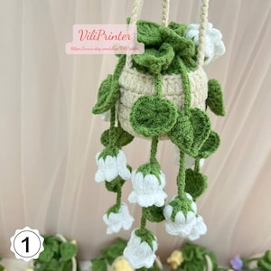 May include: Crocheted hanging planter in cream and green, featuring white flower accents. The planter has a cream-colored pot with green leaves and white flowers cascading downwards. The text "ViliPrinter" is visible on the top left.