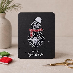 May include: A black rectangular greeting card featuring a string art snowman design made with white thread. The snowman wears a white paper hat and a red and white striped paper scarf. The text LET IT SNOW is printed in white at the bottom.