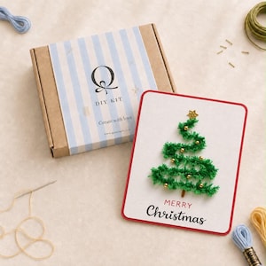 Può includere: Una scatola rettangolare in cartone per kit fai-da-te con un'etichetta a righe azzurre e bianche che riporta la lettera Q, il testo DIY KIT e Create with love. Accanto, un biglietto d'auguri bianco con bordo rosso mostra un albero di Natale.