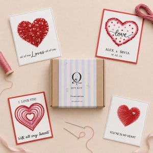 Könnte beinhalten: Ein DIY-Set-Karton mit einem hellblau-weiß gestreiften Etikett mit dem Buchstaben Q und dem Text DIY KIT Create with love, umgeben von vier Grußkarten mit herzförmigen Fadenkunst-Designs, zwei Nähnadeln und rosa Garn.