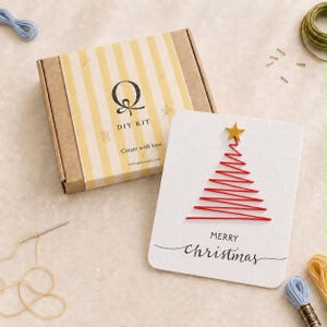 Può includere: Un kit fai-da-te in una scatola di cartone marrone a righe gialle con il testo Q DIY KIT Create with love, accanto a un biglietto bianco con un albero di Natale ricamato in rosso, una stella glitterata dorata e il testo MERRY christmas.