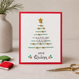 May include: A white greeting card with a red border featuring a stitched green thread Christmas tree decorated with gold beads and a gold star. The text MERRY XMAS is spelled out with white letter beads, and the card is signed LOVE QUINN.