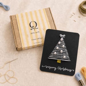 Może przedstawiać: Zestaw DIY w kartonowym pudełku w żółte paski z napisem Q DIY KIT Create with love, obok czarnej kartki z białym haftowanym motywem choinki z perłowymi akcentami oraz białym napisem Merry Christmas u dołu kartki.