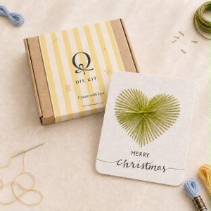 Puede incluir: Un kit de manualidades en una caja de cartón marrón con rayas amarillas y blancas, con el texto Q DIY KIT Create with love. Al lado hay una tarjeta blanca con un corazón de arte con hilos verde y el texto MERRY christmas, con hilo suelto.