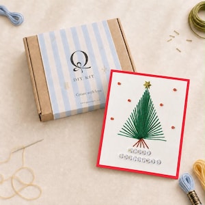 Peut inclure: Un kit DIY dans une boîte en carton à rayures bleues et blanches, avec le texte Q DIY KIT Create with love. À côté se trouve une carte de vœux blanche à bordure rouge, un sapin de Noël brodé en vert et le texte MERRY CHRISTMAS.