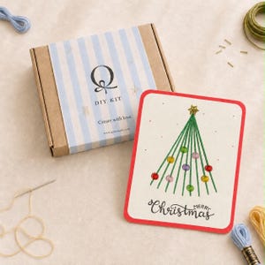 Könnte beinhalten: Ein rechteckiger DIY-Set-Karton aus Pappe mit einem hellblau-weiß gestreiften Etikett, das den Buchstaben Q, den Text DIY KIT und Create with love zeigt. Daneben liegt eine weiße Karte mit einem grünen gestickten Weihnachtsbaum und Merry Christmas.