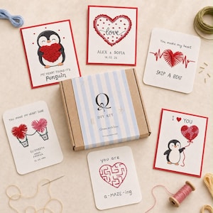 May include: A DIY craft kit box with a blue and white striped label featuring the letter Q, surrounded by six greeting cards with embroidered heart designs, including penguins, a heartbeat line, hot air balloons, and a maze, plus embroidery thread.