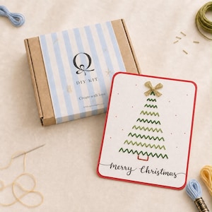 Op de afbeelding: Een doe-het-zelfpakket in een bruine kartonnen doos met een lichtblauw gestreept label met de letter Q en de tekst DIY KIT Create with love. Daarnaast ligt een witte kaart met een rode rand, een groene geborduurde kerstboom en Merry Christmas.
